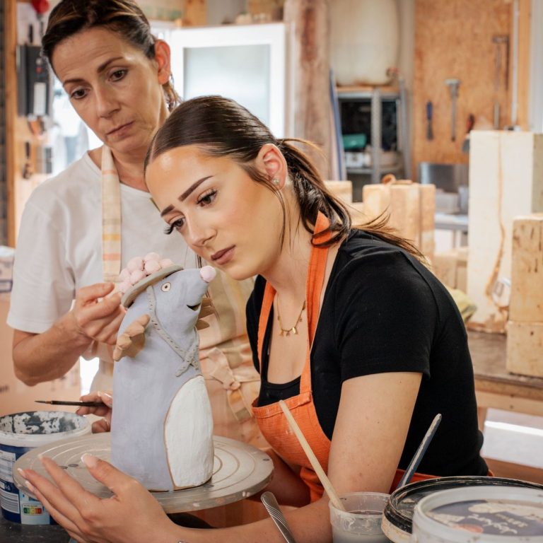 Bei der Arbeit Zwei Frauen modellieren eine Skulptur in einer Werkstatt. Eine beobachtet aufmerksam.